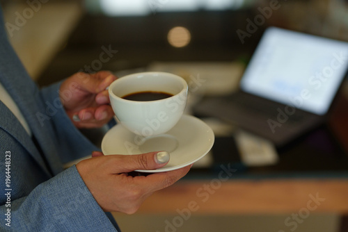 Fotografija Hands holding a white cup of black coffee and saucer, a laptop with a bright scr
