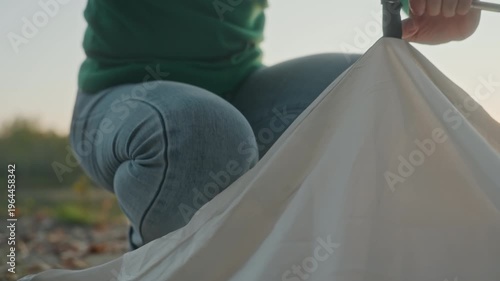 Closeup hands securing tent pole at lakeside sunset, detailed fabric and rope work, fasten clips and stakes with careful precision, jeans and green sweater visible, calm evening gear setup
