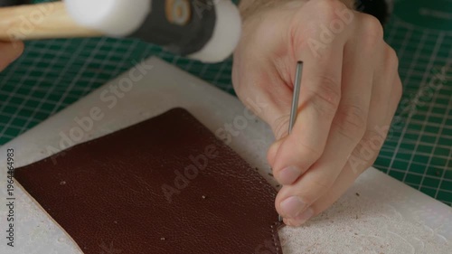A close-up cinematic shot showing a leather craftsman using a small metal hammer and a round punch to create a series of perfect holes in a strip of natural brown leather.