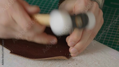 A close-up cinematic shot showing a leather craftsman using a small metal hammer and a round punch to create a series of perfect holes in a strip of natural brown leather.