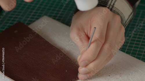 A close-up cinematic shot showing a leather craftsman using a small metal hammer and a round punch to create a series of perfect holes in a strip of natural brown leather.