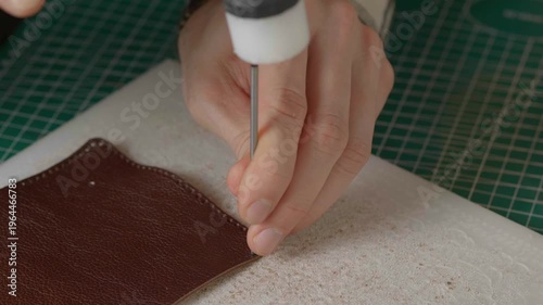 A close-up cinematic shot showing a leather craftsman using a small metal hammer and a round punch to create a series of perfect holes in a strip of natural brown leather.