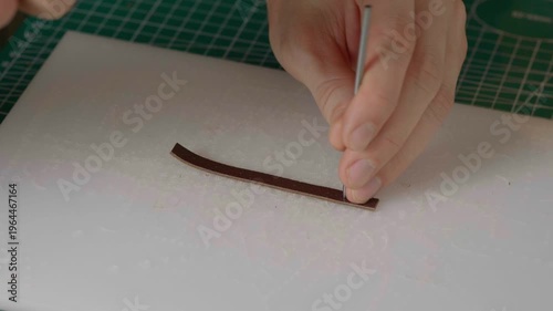 A close-up cinematic shot showing a leather craftsman using a small metal hammer and a round punch to create a series of perfect holes in a strip of natural brown leather.