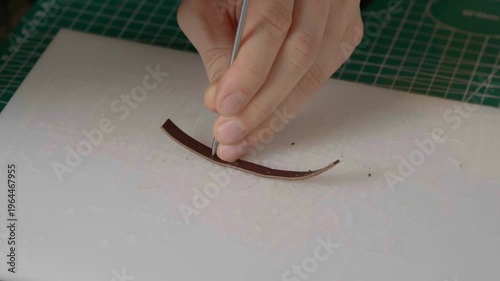 A close-up cinematic shot showing a leather craftsman using a small metal hammer and a round punch to create a series of perfect holes in a strip of natural brown leather.