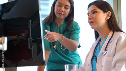 Two female medical professionals, a doctor and an assistant, are in a bright and modern hospital setting, intently reviewing patient results on a computer screen. 