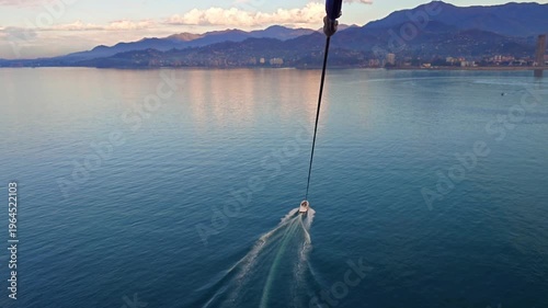 Aerial view of a speedboat pulling a parasail over calm sea, leaving wake trails. Scenic coastline with mountains and city in soft evening light, travel and adventure concept.