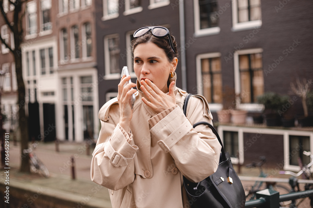 Fototapeta premium Surprised young woman in a beige trench coat covering her mouth while talking on a smartphone on a city street. Female traveler looking aside with a shocked expression on a European background.