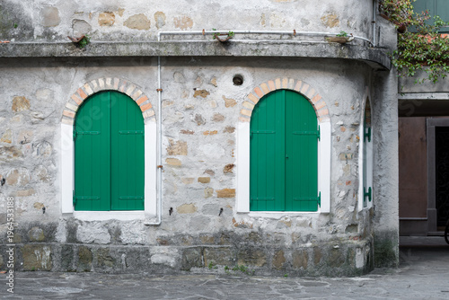 Historic stone wall with arched windows and green wooden shutters in Grado, Italy. Rustic Mediterranean architecture in the old town alley, traditional Italian building facade with brick details.