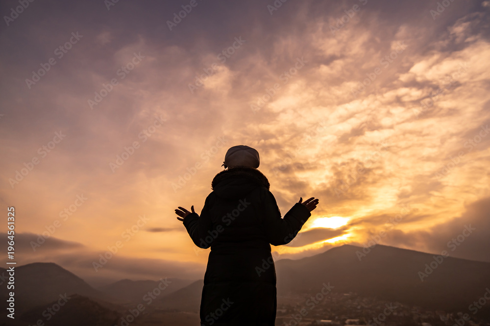Fototapeta premium Woman sunset silhouette stands on mountain with arms open toward dramatic golden sky