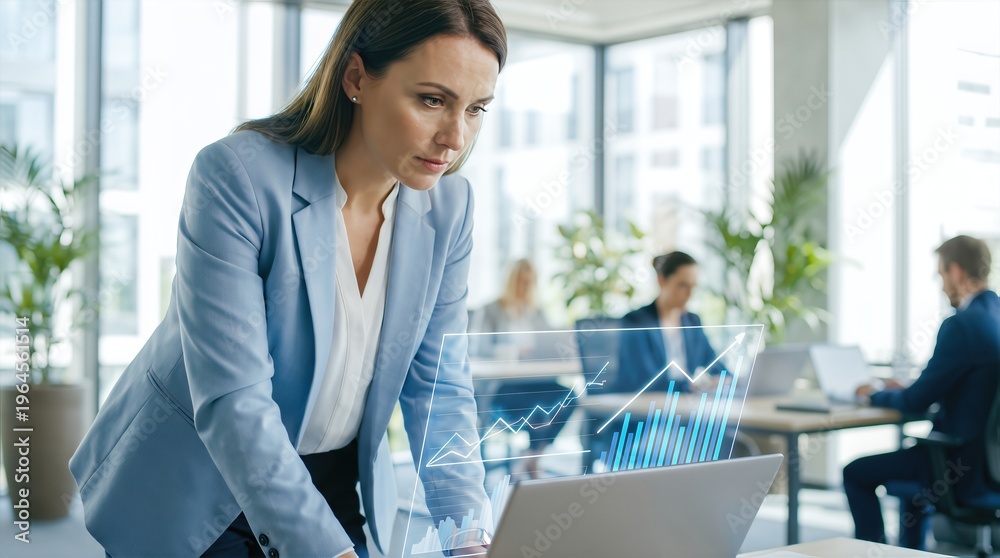 Obraz premium Businesswoman in a light blue blazer analyzes financial data on a laptop with holographic graphs in a modern office environment filled with plants and colleagues