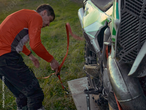 Man secures dirt bike with ratchet strap on motorcycle trailer. Athlete preparing off-road motorcycle for transport. Extreme sports hobbyist loading gear for travel to competition venue.