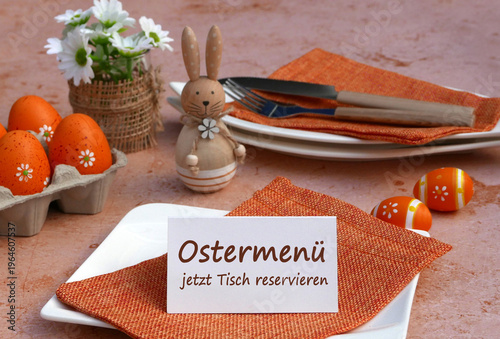 Tischkarte mit dem Text Ostermenü jetzt reservieren neben orangefarbener Osterdekoration und Frühlingsblumen auf einem gedecktem Tisch.