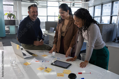 Diverse coworkers leaning and reviewing printed charts with laptop, tablet, sticky notes at office