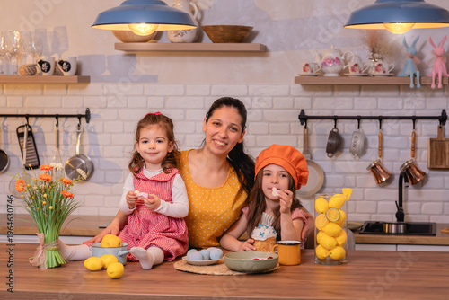 happy mother with little daughters celebrating easter in cozy kitchen with flowers sweets and painted eggs, warm spring family moment together