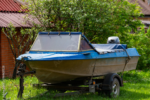 Blue Motorboat On Trailer Parked In Backyard With Outboard Engine And Weathered Hull
