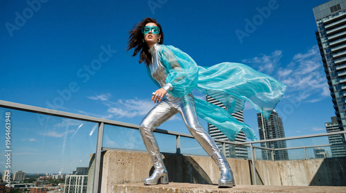Model poses in silver outfit with blue cape on rooftop against city skyline under bright sky