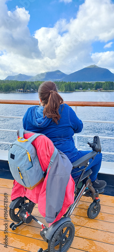 Young woman in a wheelchair sits looking out over the ocean from the deck of a cruise ship, travelling in Alaska, enjoying the freedom from disability the mobility aid allows her to travel.