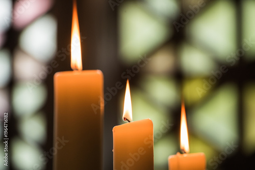 Three burning candles in a church with colorful stained glass window bokeh background
