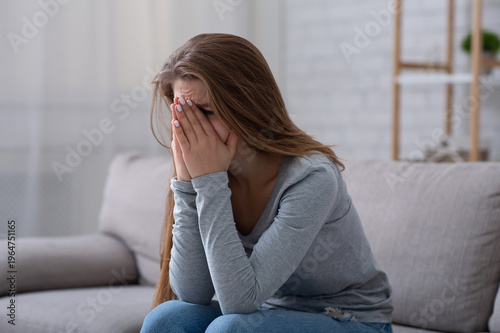 Fotografie Young woman burying her face in hands and crying, feeling depressed or desperate on sofa indoors