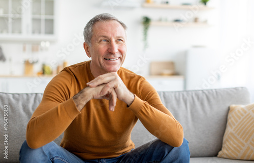 фотографія Portrait of cheerful casual senior man smiling and looking away, sitting on sofa couch in living room