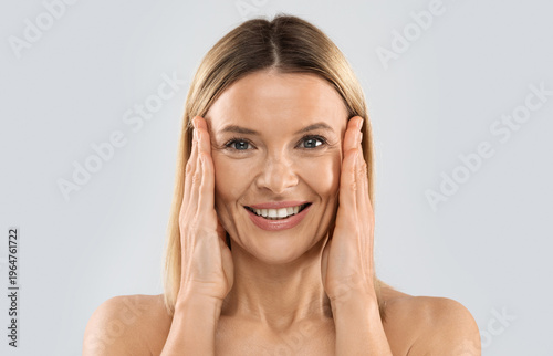 Closeup of happy pretty nude middle aged blonde woman smiling at camera and touching face, posing on grey studio background, pulling face skin, copy space. Plastic surgery, face-lift concept