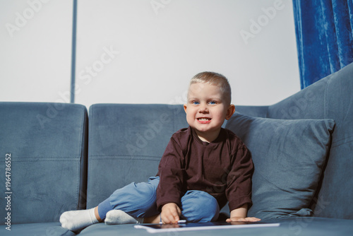 Obraz na płótnie Child sits on a couch and plays with a tablet in a bright room with blue curtain
