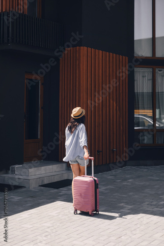 Woman tourist with pink luggage walking to modern hotel entrance. Female traveler arriving at rental apartment during summer vacation trip. Rear view of person moving into new house.
