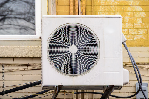 Single mini-split air conditioner condenser unit mounted on exterior wall of yellow brick building, pipes and wiring visible.