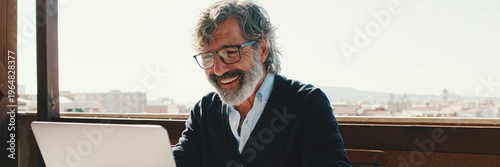 Happy middle aged man communicates remotely using laptop. Working from home office. Panoramic