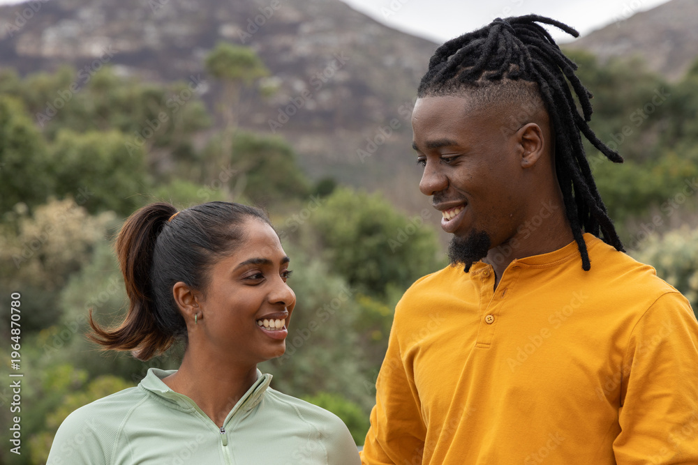 Fototapeta premium Diverse couple smiling and facing each other while standing on trail in hills wearing activewear