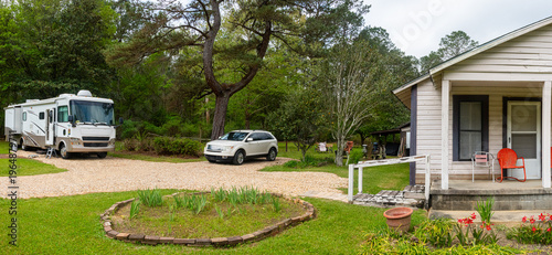 A camper van is parked in a rustic setting beside an old wooden house. The scene is framed by vibrant garden flowers and a stone porch.