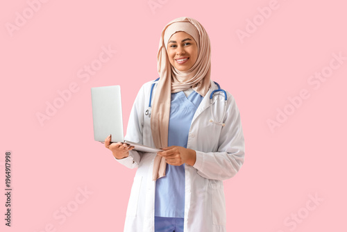 Female Muslim medical student with laptop on pink background