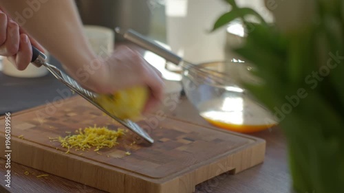 Hand grates lemon on fine metal grater with fresh zest on wooden cutting board. Concept of cooking and baking ingredient preparation.