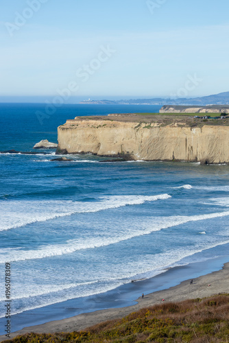 Scenic view of coastal cliffs and sandy beach along California Highway 1. Blue Pacific Ocean waves roll toward rugged shoreline under clear sky on the California coast