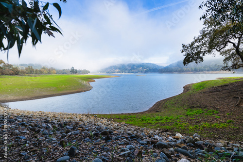 Foggy morning over a calm lake with curved shoreline, rocky foreground and eucalyptus trees. Soft mist covers distant hills creating a peaceful natural landscape scene