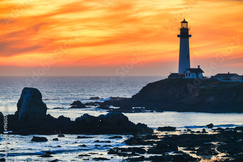 Pigeon Point Lighthouse glowing at vibrant sunset over rocky Pacific Ocean shoreline in California, with dramatic orange sky, coastal cliffs and textured seascape