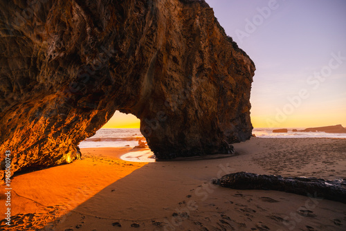 Natural sea arch at Hole in the Wall Beach in California during sunset, warm golden light illuminating rugged coastal rock, ocean waves and smooth sand, creating dramatic seaside landscape