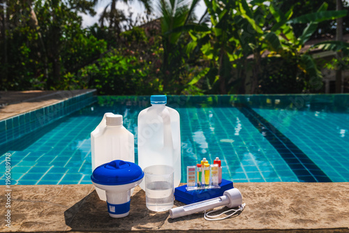 Swimming pool maintenance chemicals and water testing kit with pH and chlorine test tubes, measuring cup and dispenser arranged on pool edge, outdoor pool water in background