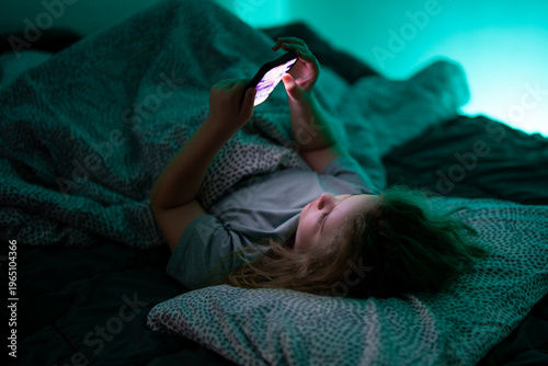 Child holding smartphone under blanket light. Kid using technology during bedtime routine. Child watching video on phone at night. Kid scrolling content on mobile device in bed.
