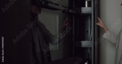 A person is seen opening a sleek, modern glass cabinet in a dimly lit room, creating a reflective and mysterious atmosphere. Person Opening a Modern Glass Cabinet in a Dimly Lit Room