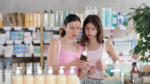Woman with teen girl choose buy expectorant syrup at pharmacy, compare with other good. Client check for absence of harmful substances, view product in details. 