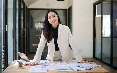 Confident businesswoman standing at a desk with financial reports and charts, smiling in a modern office workspace.