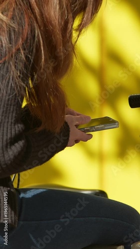 On the city streets, a Japanese woman, near a bicycle, engages in a text conversation on her smartphone with a yellow wall background. An Asian lady on a bike uses her phone on the street