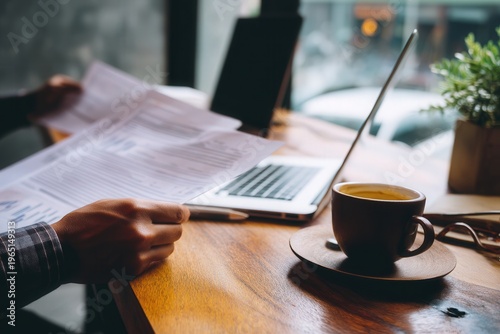 Person reviewing financial documents at a wooden desk with laptop and coffee cup in a cozy cafe workspace.