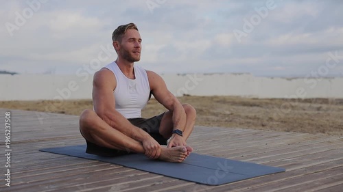 Young happy strong sporty athletic fit sportsman man wear sports clothes sitting on rubber mat rest relax warm up train at sunrise sun dawn over sea beach outdoor on pier seaside in summer day morning