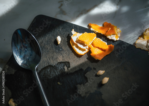 POV close up of orange peel scraps and seeds on dark plate with spoon under dramatic natural light creating contrast and texture i