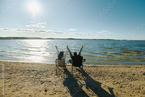 Two people relax on chairs by the lake under the sun enjoying the warm weather while watching the water