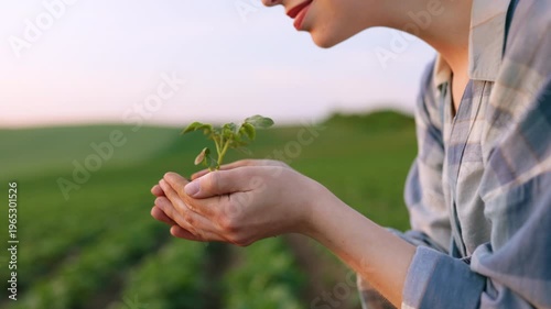A young woman gently holds a small green seedling in her cupped hands, smelling its fresh scent in a field at sunset
