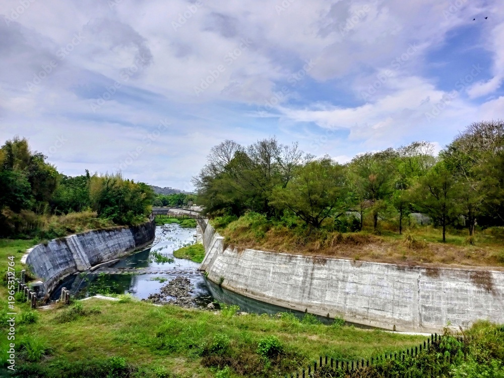 Fototapeta premium Abandoned Concrete Structure by River: The remnants of a concrete structure, perhaps a dam or water channel, stand weathered and worn along the bank of a flowing river.