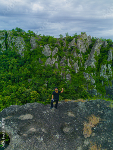 Aerial photo of a male adventurer on a rocky mountaintop. Drone photo of a solo traveler on a pristine & beautiful ancient rocky hilltop with a backdrop of other rocky mountains covered in green trees